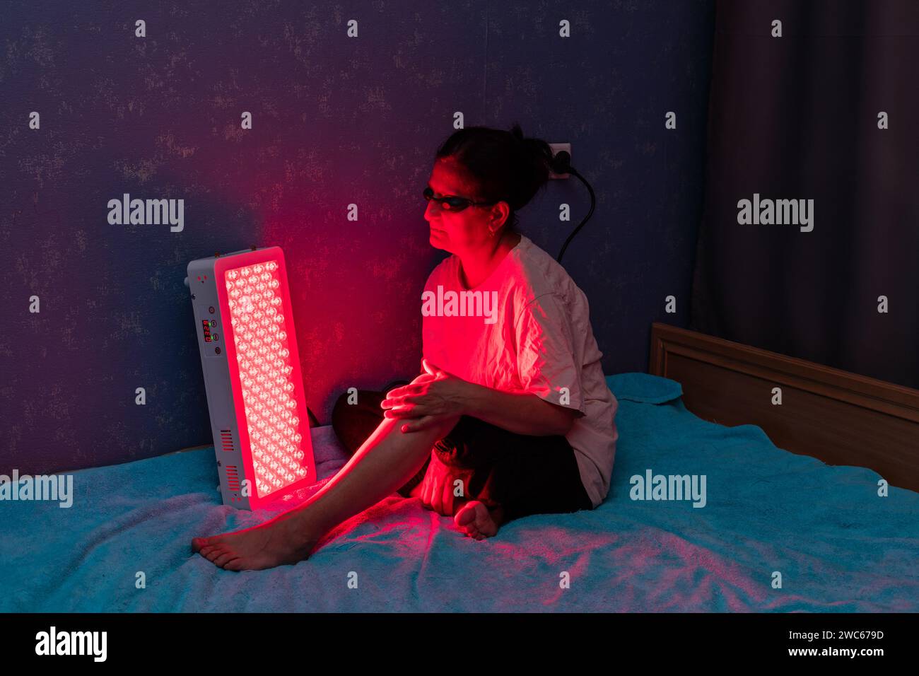 Femme âgée traitant la jambe douloureuse avec le panneau de thérapie de lumière rouge et portant une protection oculaire à la maison sur le lit Banque D'Images