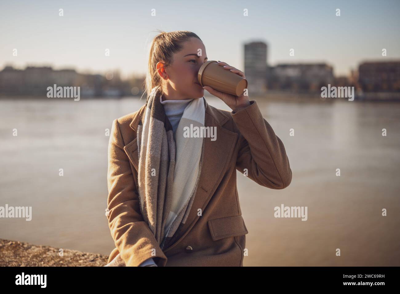 Belle femme dans des vêtements chauds aime boire du café et se reposer au bord de la rivière sur un jour d'hiver ensoleillé.image tonique. Banque D'Images