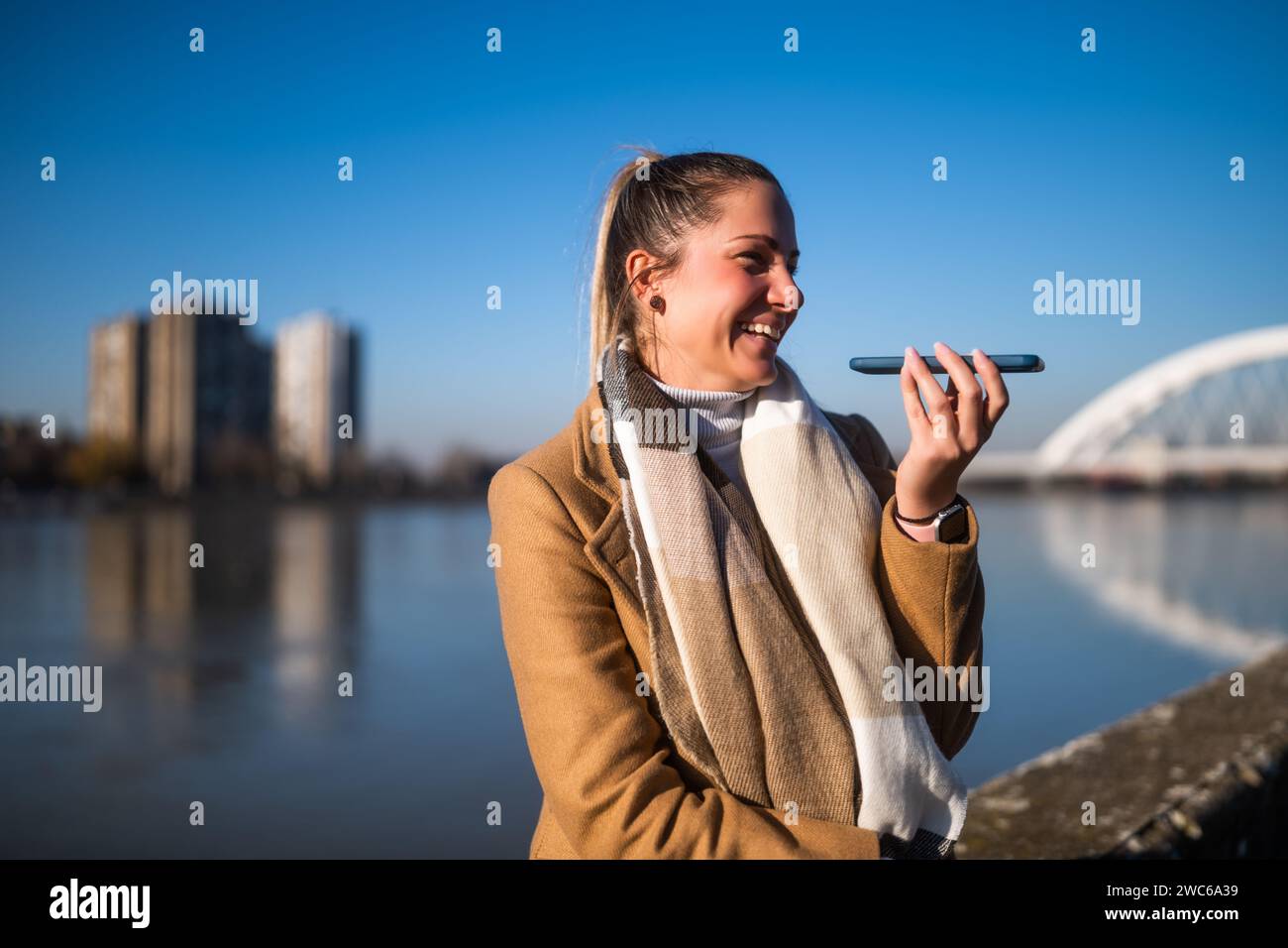 Belle femme en vêtements chauds parlant sur un téléphone portable et aime se reposer au bord de la rivière par une journée ensoleillée d'hiver. Banque D'Images