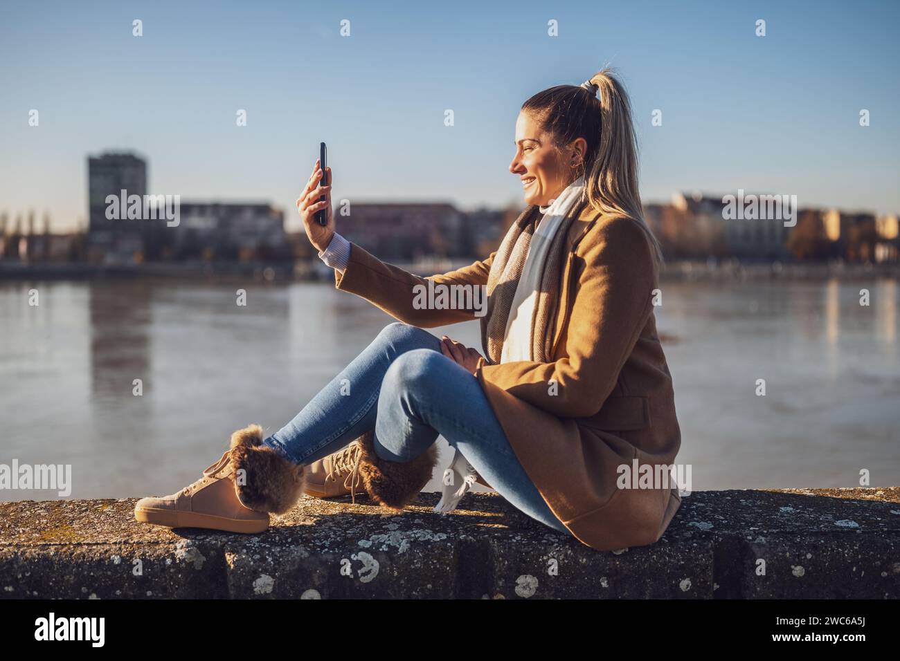 Belle femme dans des vêtements chauds prenant selfie avec téléphone et aime se reposer au bord de la rivière sur un jour d'hiver ensoleillé.image tonique. Banque D'Images