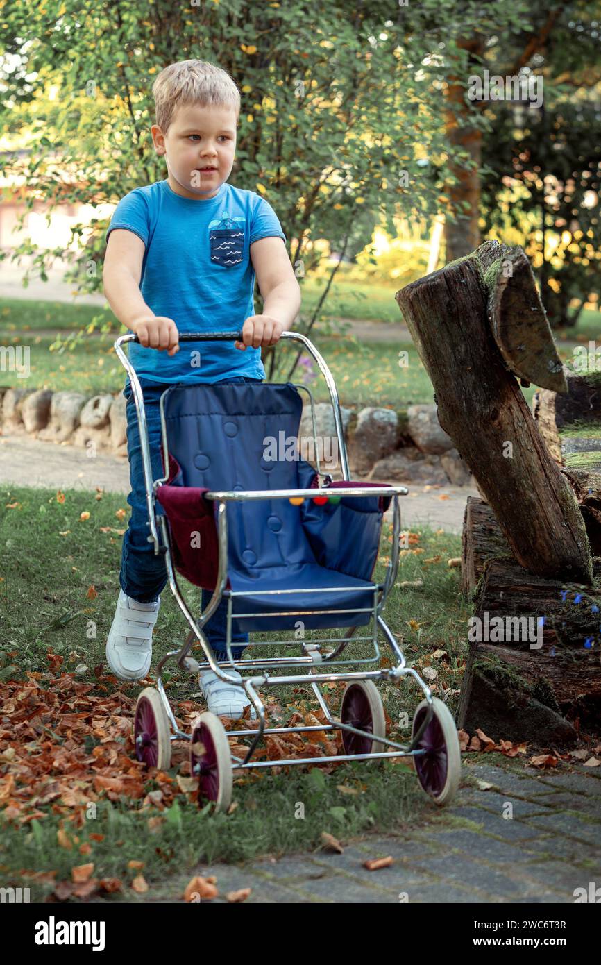 Un petit garçon jouant avec des jouets pour les filles, il pousse un chariot de poupée dans un parc d'automne. Banque D'Images