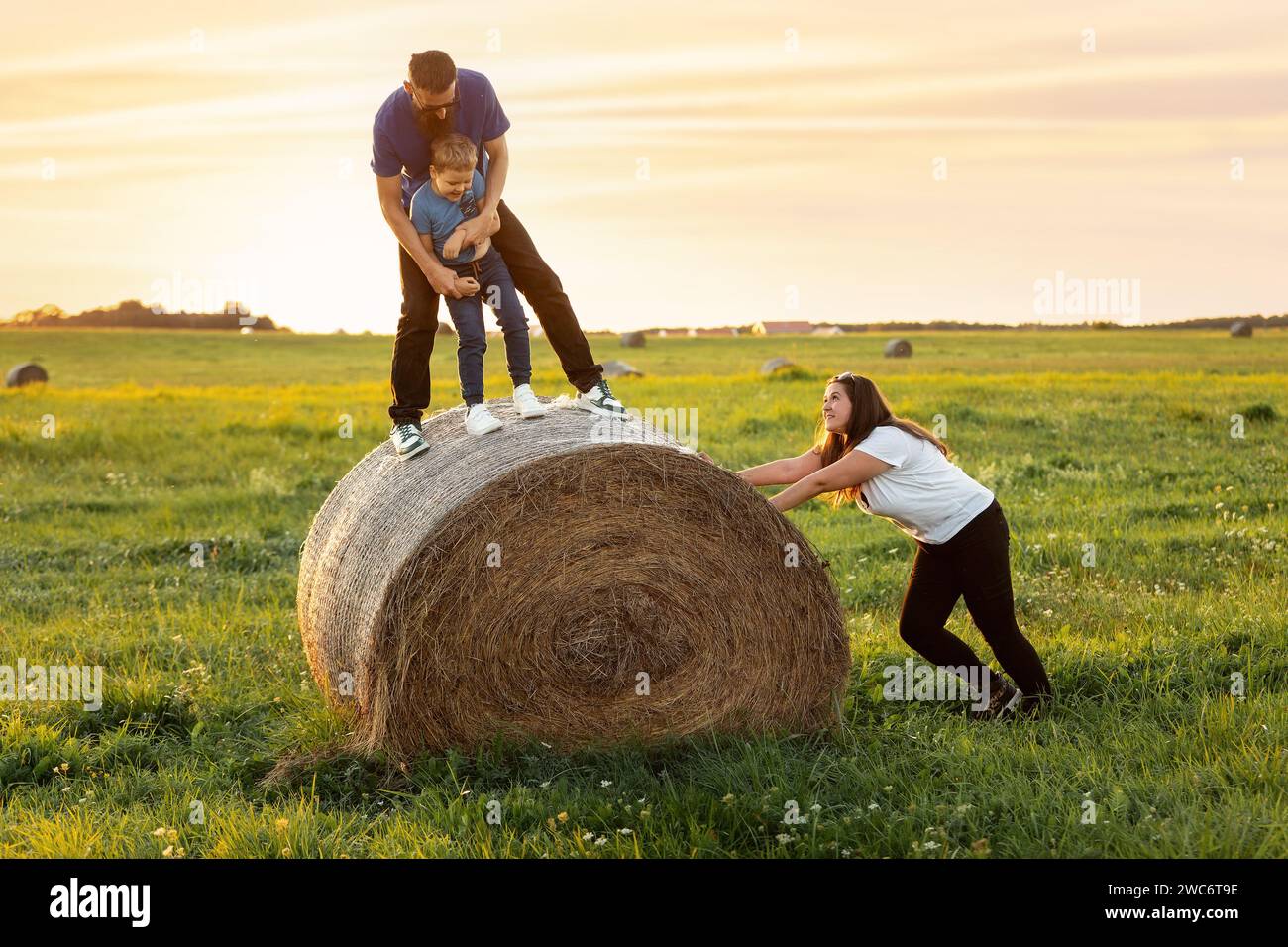 Jeux de famille amusants sur une pelouse rurale à l'extérieur le soir au coucher du soleil. Une mère pousse un rouleau de foin et veut que son mari et son fils tombent. Un fami harmonieux Banque D'Images