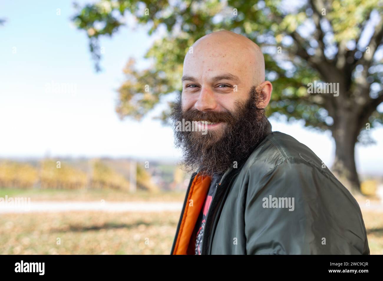 Un homme avec une longue barbe abîmée souriant par temps ensoleillé. Il est en plein air, chauve et porte un blouson d'aviateur Banque D'Images
