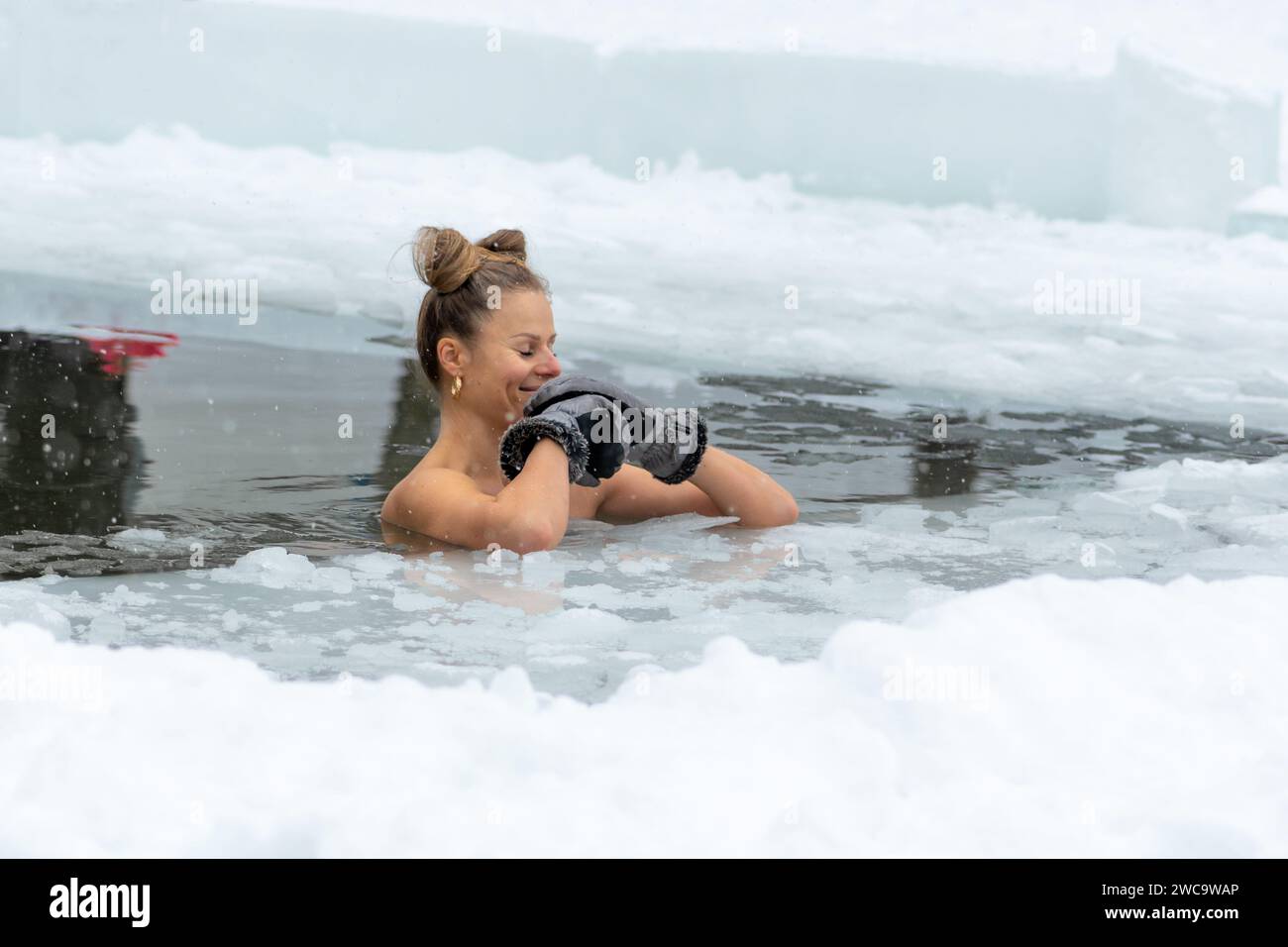Belle fille aux cheveux blonds portant des gants de bain de glace dans l'eau froide d'un lac gelé. Méthode Wim Hof, thérapie par le froid Banque D'Images