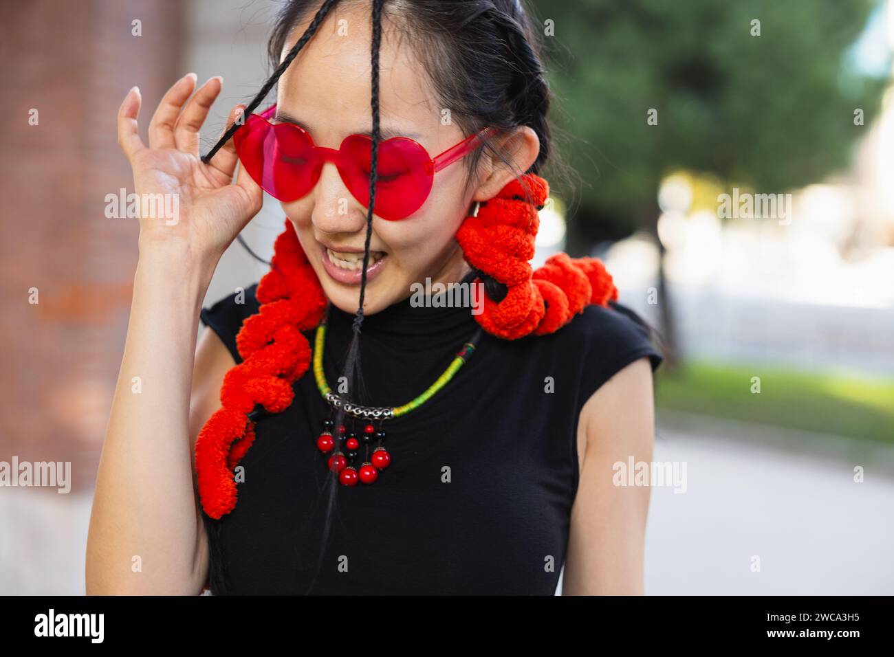 'Une photo franche d'une danseuse de rue souriante tout en ajustant ses lunettes de soleil rouges en forme de cœur, ornées d'accessoires rouges éclatants. Banque D'Images