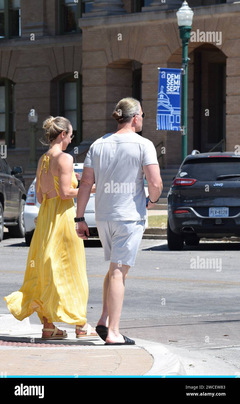 Homme millénaire avec un chignon de cheveux et femme en robe de soleil jaune attendre sur un coin de rue pour traverser une rue dans le centre-ville de Georgetown Texas - août 2023 Banque D'Images