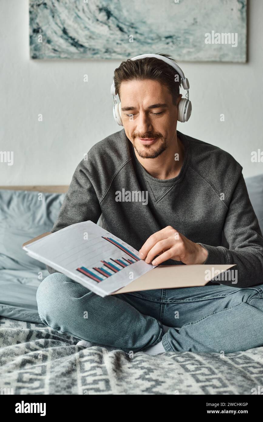 homme barbu dans des écouteurs sans fil analysant des graphiques tout en écoutant de la musique dans la chambre, le travail de la maison Banque D'Images