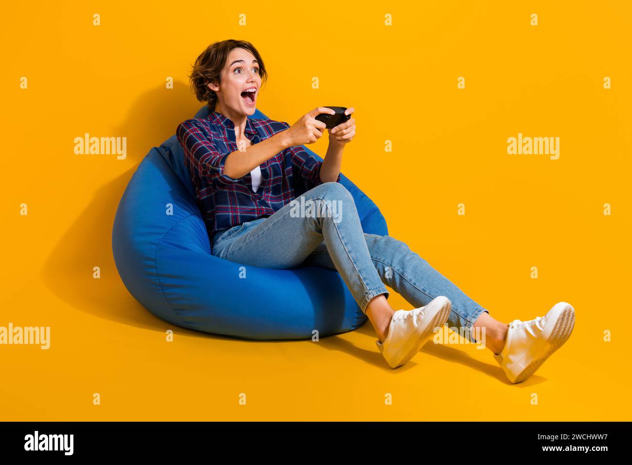 Photo grandeur nature de fille extatique porter chemisier à carreaux s'asseoir sur le pouf Hold joystick look espace vide isolé sur fond de couleur jaune Banque D'Images