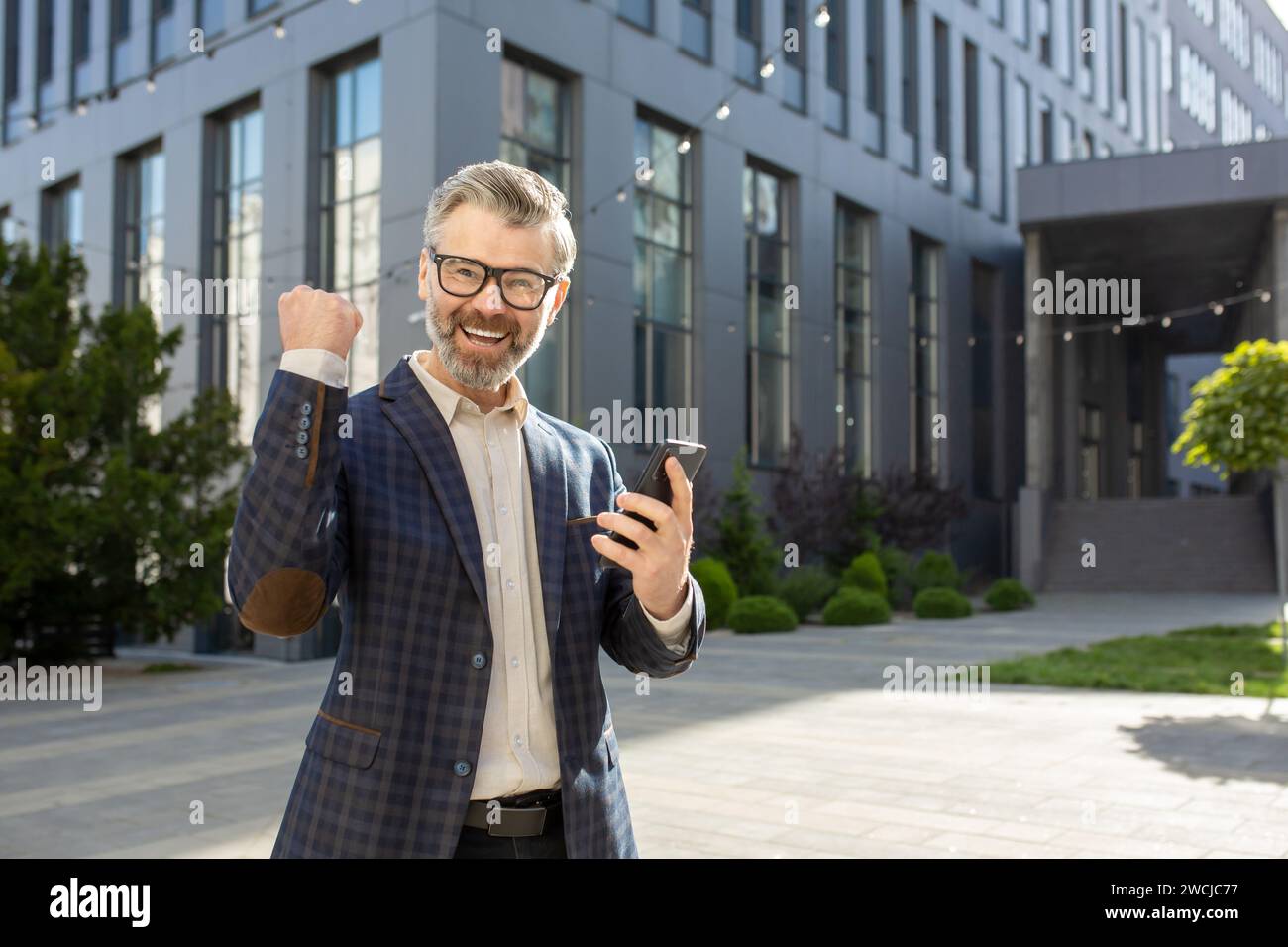 Joyeux cadre dirigeant montrant un geste de victoire avec un poing levé tout en tenant un smartphone, debout à l'extérieur d'un bureau moderne. Banque D'Images