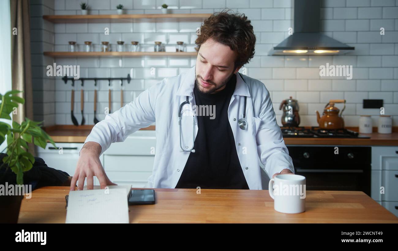 Jeune médecin masculin professionnel porter un uniforme blanc avec stéthoscope ayant la conférence téléphonique vidéo, discuter en ligne avec le patient, regarder ses notes Banque D'Images