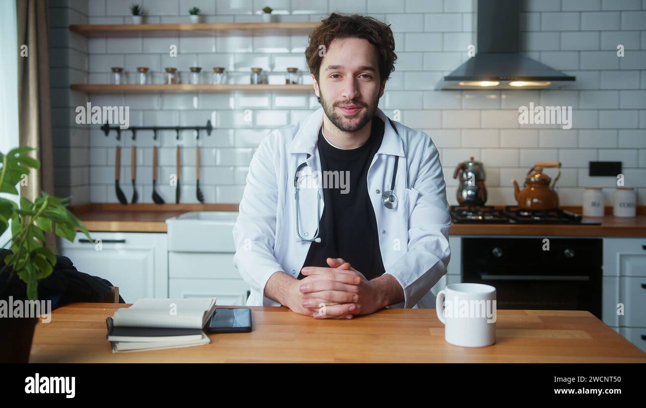 Jeune professionnel des cheveux bouclés médecin masculin porter un uniforme blanc avec stéthoscope ayant la conférence téléphonique vidéo, discuter en ligne avec le patient, regarder la caméra i Banque D'Images