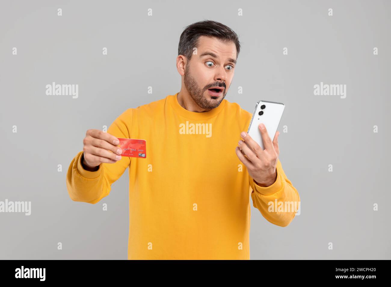 Homme choqué avec carte de crédit et smartphone sur fond gris. Problème de dette Banque D'Images