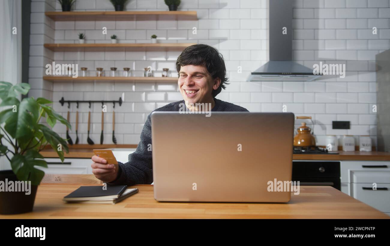 Jeune homme souriant assis dans la cuisine entrant le numéro de carte de crédit sur l'ordinateur portable pour rend sécurisé facile paiement électronique à distance à la maison Banque D'Images