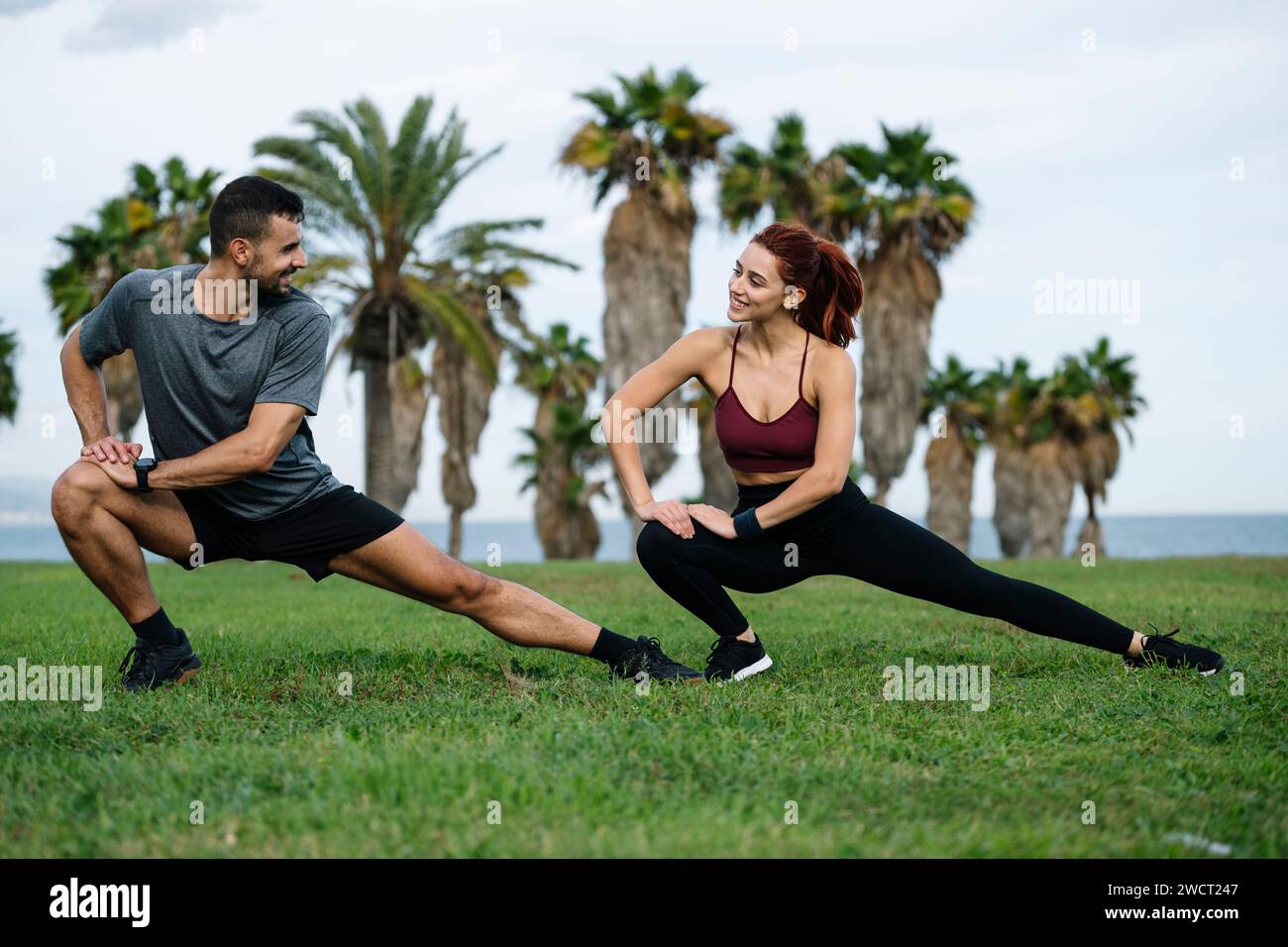 Souriant satisfait athlétique masculin et féminin assis dans l'herbe dans l'entraînement d'entraînement de sportswear à l'extérieur. Heureux jeune couple adulte étirant dans le Banque D'Images