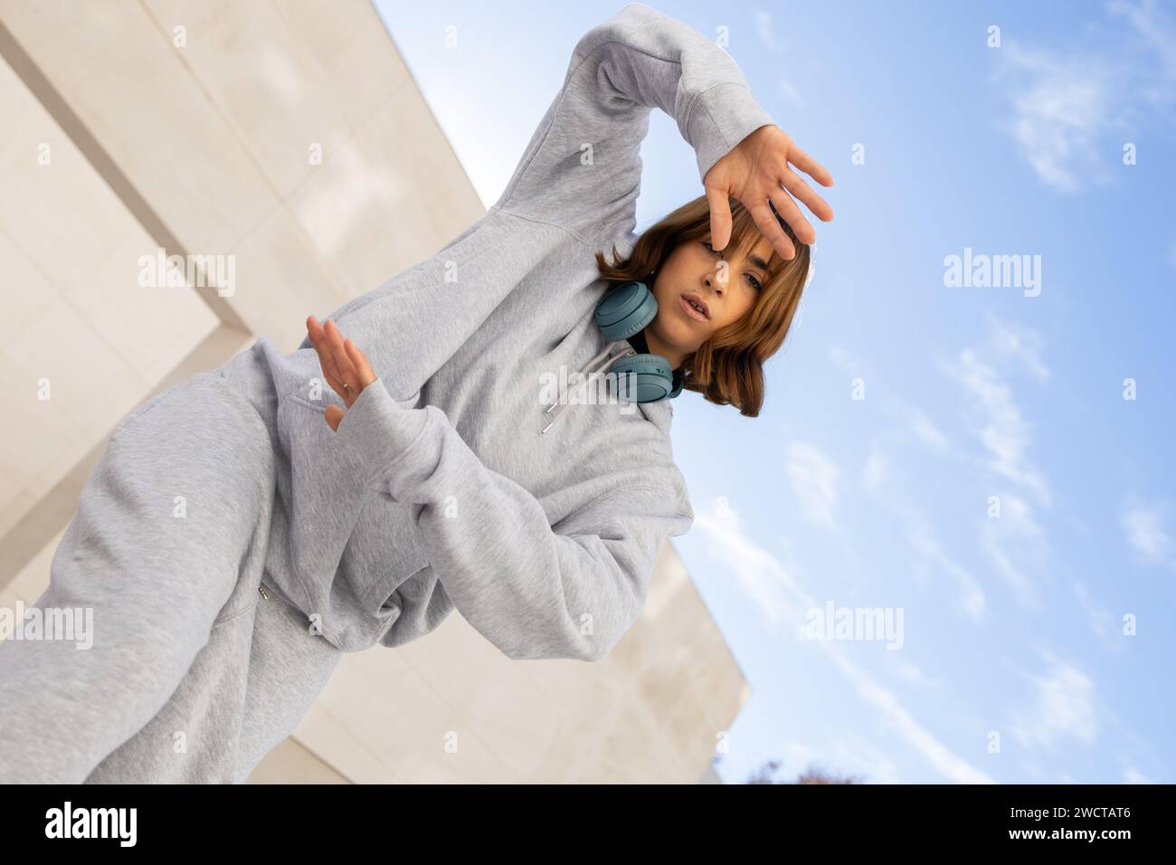 Une jeune femme prend une pose élégante dans son survêtement gris confortable, accessoirisé avec des écouteurs sans fil tendance, sous un ciel bleu clair Banque D'Images