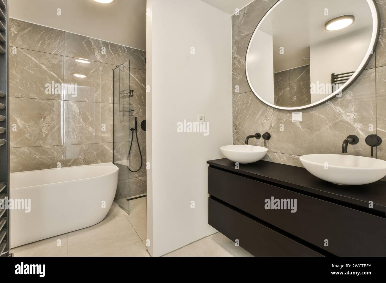 Salle de bain contemporaine avec carrelage mural en marbre poli, cabine de douche en verre sans cadre, baignoire sur pied, vasques ovales sur meuble sombre Banque D'Images