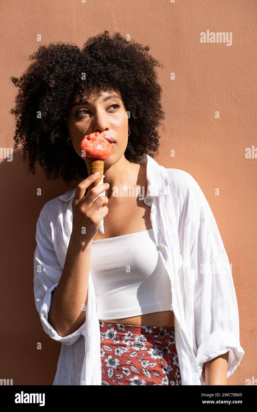 Femme noire aux cheveux frisés en blanc apprécie un cône de crème glacée fraise contre un mur chaud de pêche regardant loin Banque D'Images