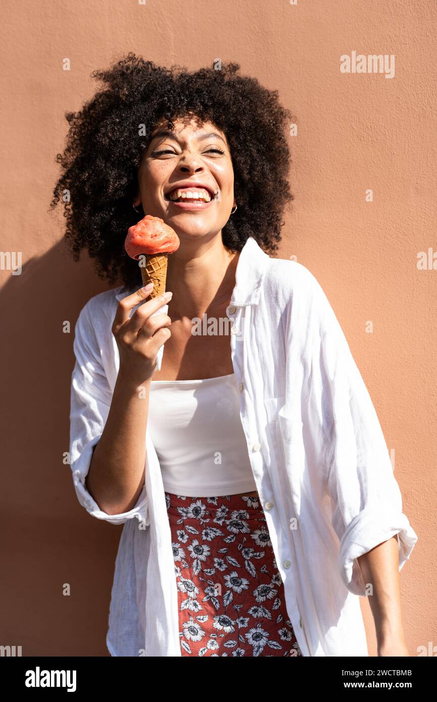 Femme noire aux cheveux frisés en blanc apprécie un cône de crème glacée fraise contre un mur chaud de pêche regardant loin Banque D'Images