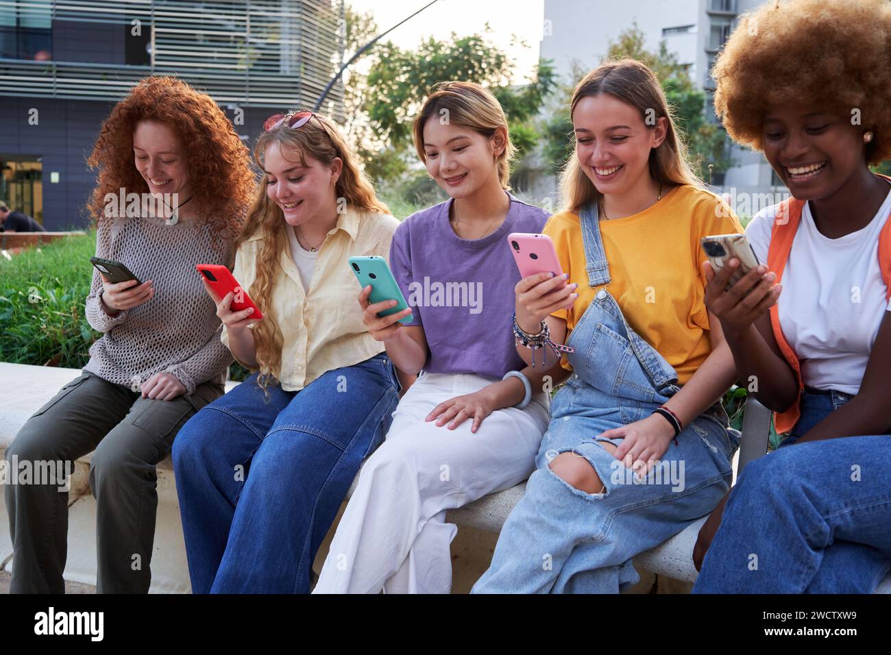 Groupe multiracial de jeunes amies appréciant et souriant en utilisant leurs téléphones assis dehors. Banque D'Images