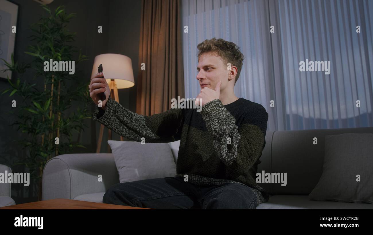 Bel homme blond assis sur le canapé ayant la conversation d'appel vidéo avec le téléphone intelligent. Partagez des nouvelles, parlez à votre famille, profitez de réunions virtuelles de chat en ligne Banque D'Images