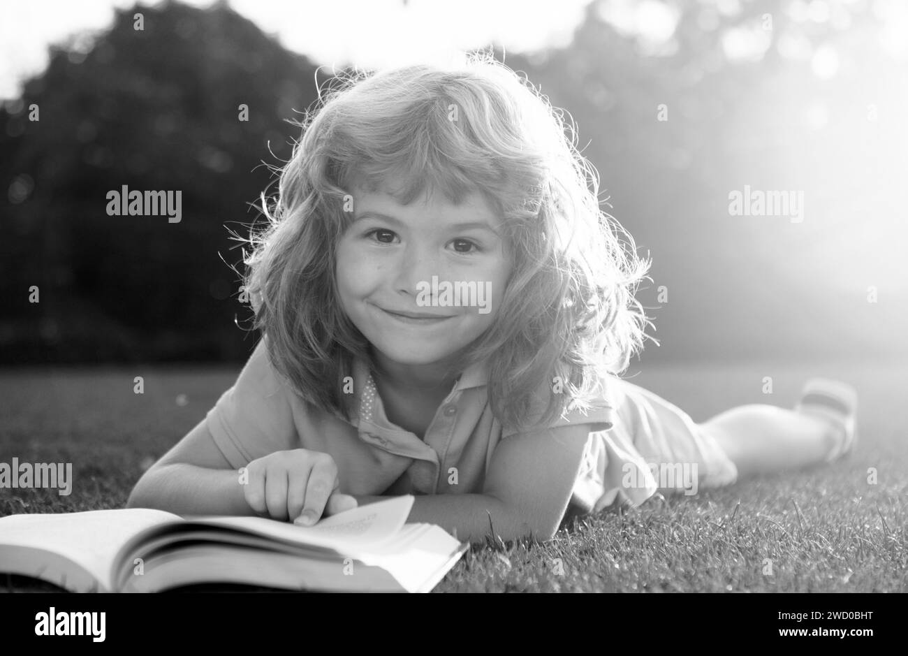 Enfant garçon lisant un livre couché sur l'herbe. Mignon petit enfant dans des vêtements décontractés lisant un livre et souriant tout en étant allongé sur l'herbe dans le parc. Banque D'Images