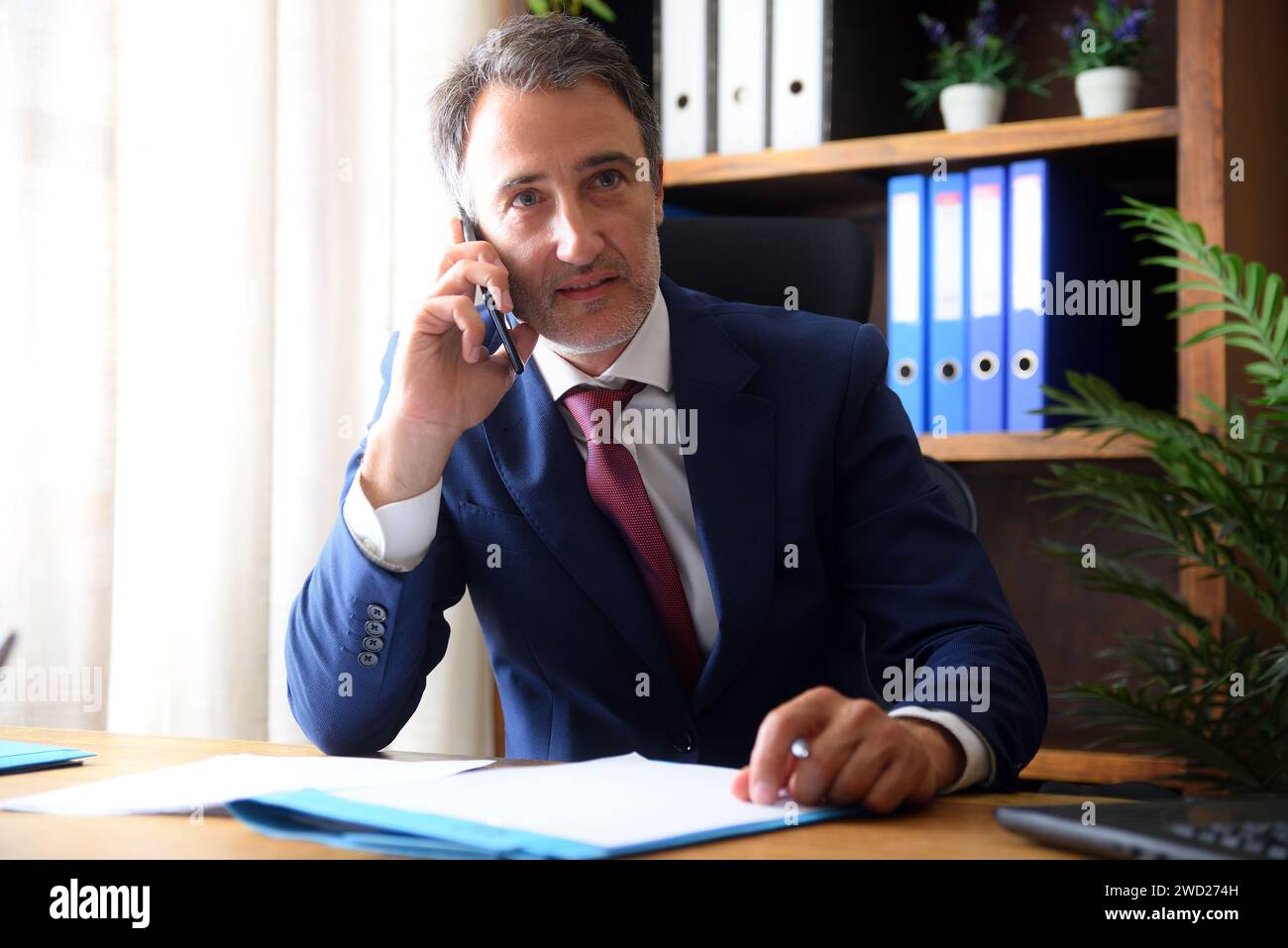 Homme d'affaires en costume bleu et cravate parlant sur le téléphone portable au bureau avec des documents sur la table et des étagères en arrière-plan Banque D'Images