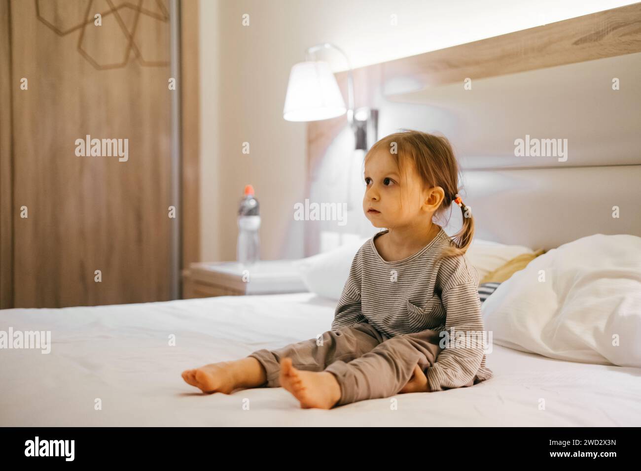Jeune fille fatiguée assise dans la chambre d'hôtel sur le lit le soir. Avant le coucher enfant triste attendant de dormir. Banque D'Images
