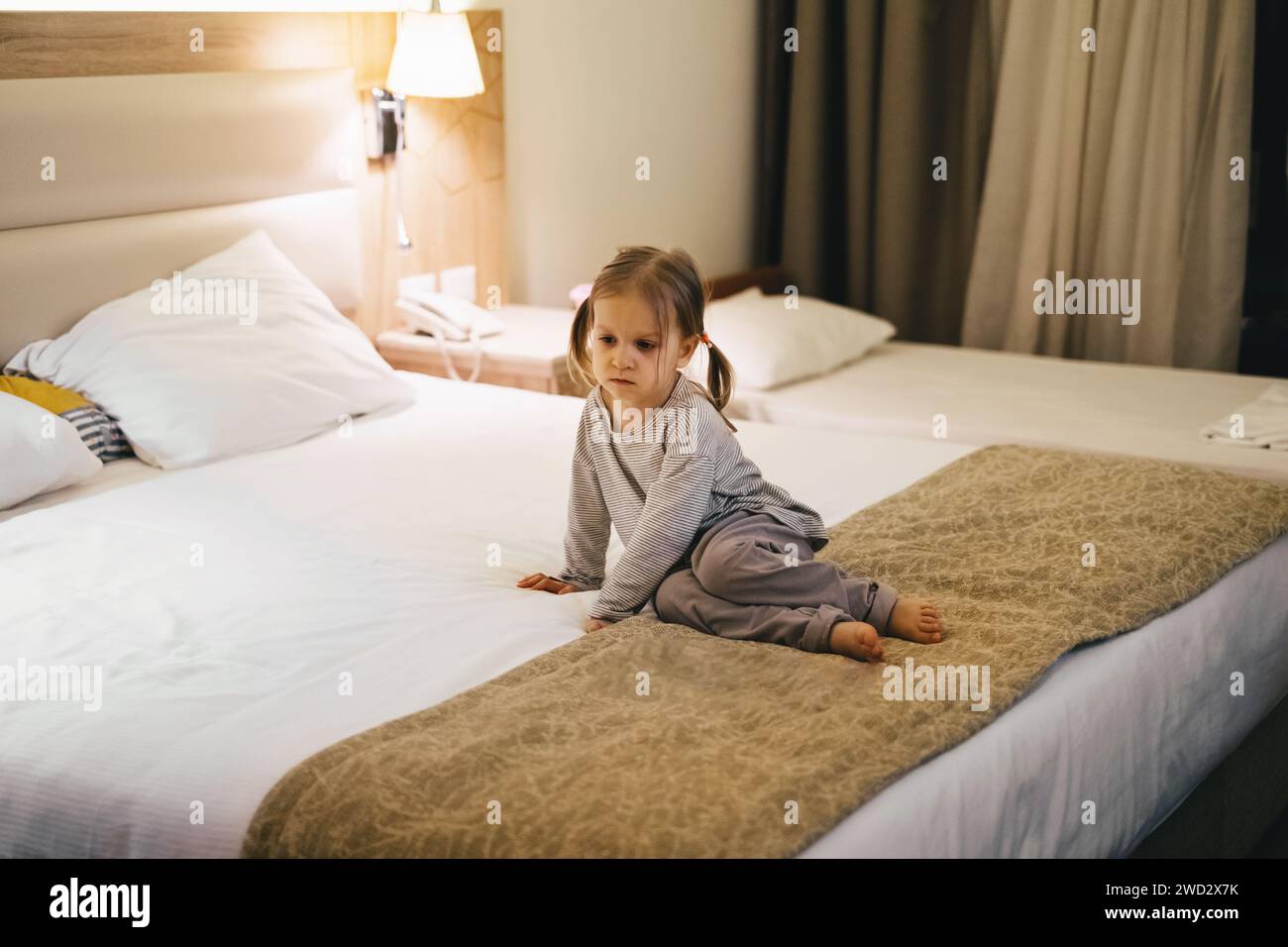 Jeune fille fatiguée assise dans la chambre d'hôtel sur le lit le soir. Avant le coucher enfant triste attendant de dormir. Banque D'Images
