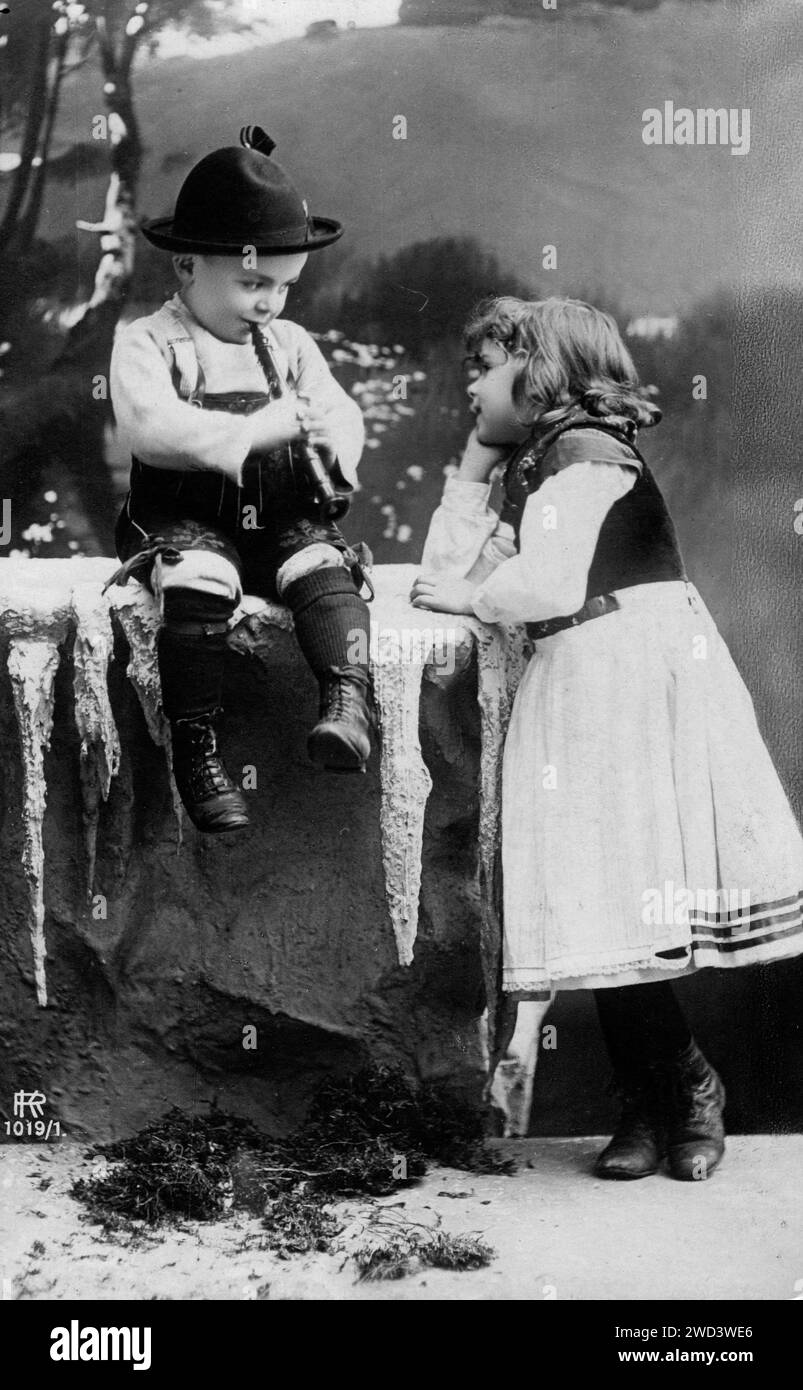Une carte postale antique montre un garçon en vêtements traditionnels et un chapeau jouant de la pipe à une fille qui se tient à proximité et l'écoute, en posant sa tête dessus Banque D'Images