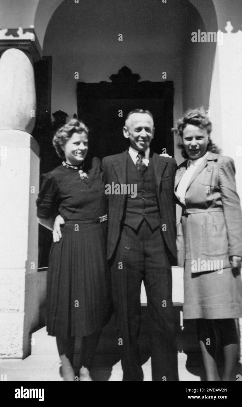 Une photo antique montre un homme d'âge moyen en costume d'affaires se tient debout avec deux femmes élégantes, apparemment sa femme et sa fille, au seuil d'un pri Banque D'Images