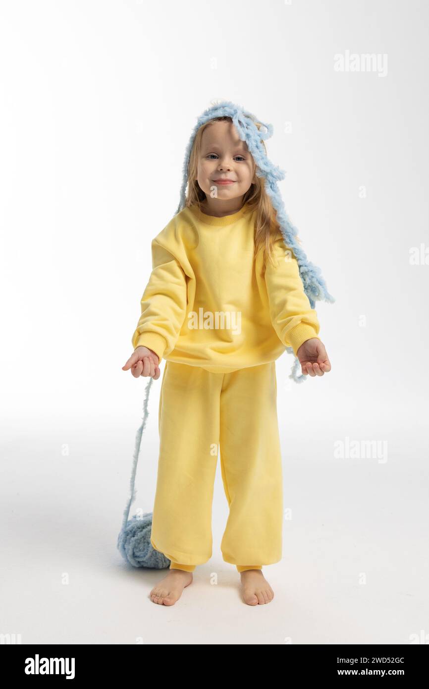 Jolie petite fille en vêtements jaunes joue avec boule de fil bleu. Modèle enfant avec de longs cheveux blonds assis dans le studio sur fond clair. Photo de haute qualité Banque D'Images