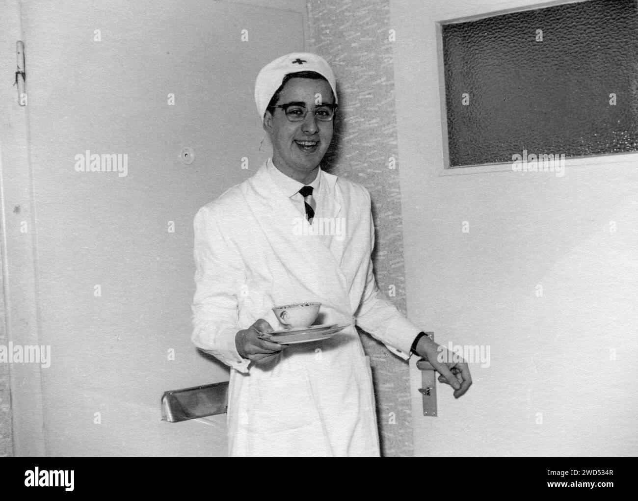 Une photo antique avec montre un jeune homme dans un manteau blanc, un travailleur médical, et un chapeau avec une croix rouge se tient près de la porte et tient une tasse de thé ou c Banque D'Images