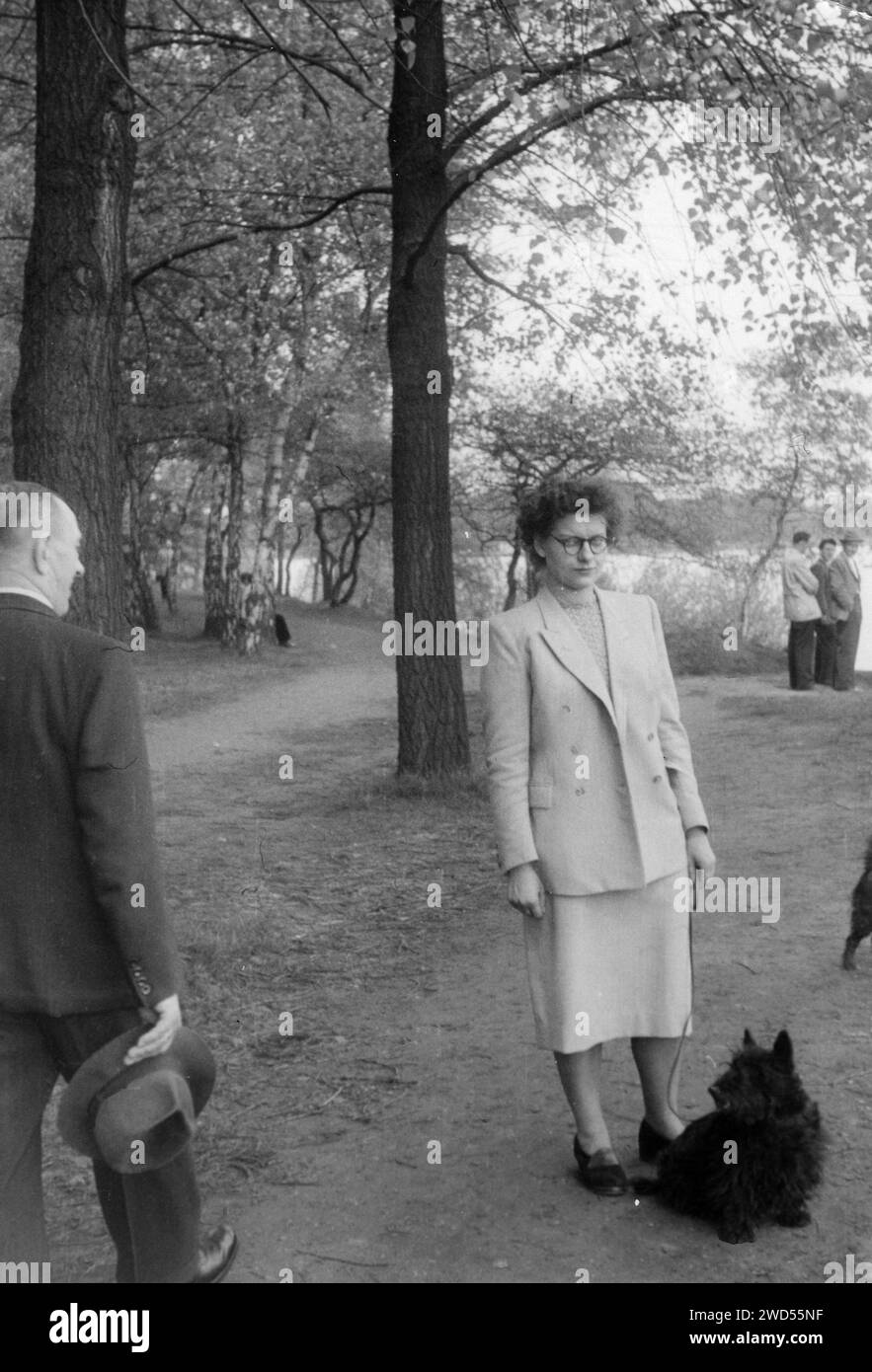 Une photo antique avec montre une femme dans un costume minable avec un chien près de ses pieds posant dans le parc, un homme âgé dans un costume formel passe devant eux, lui Banque D'Images