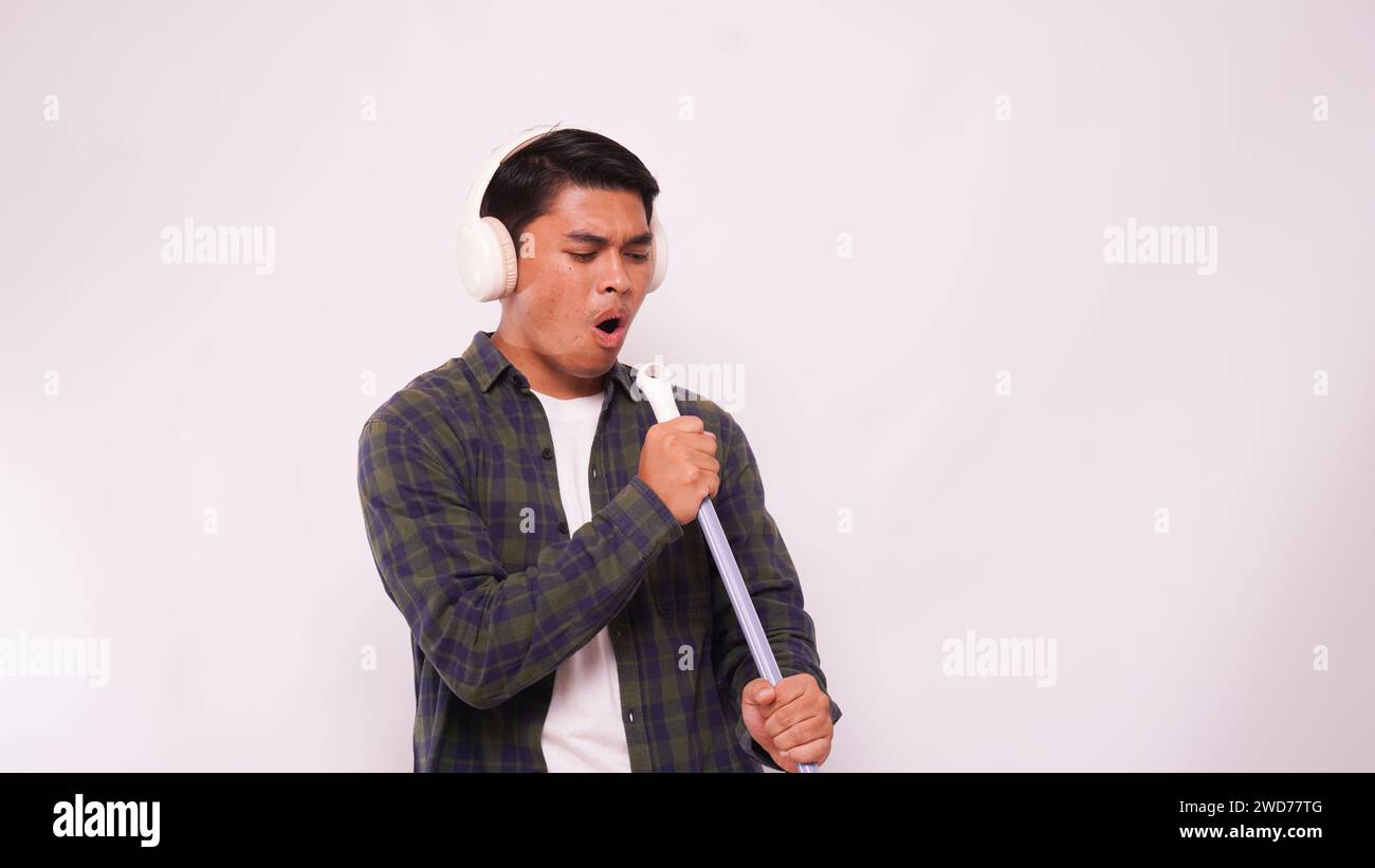 Homme asiatique écoutant de la musique avec un casque et chantant sur un balai sur fond blanc Banque D'Images