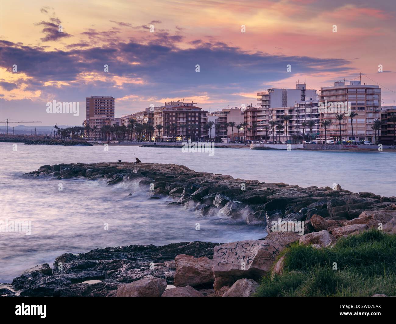 Torrevieja, Alicante, Espagne. Derrière un pêcheur solitaire sur le brise-lames, les lumières de la ville de Torrevieja clignotent alors que le soleil se couche sur la Méditerranée Banque D'Images