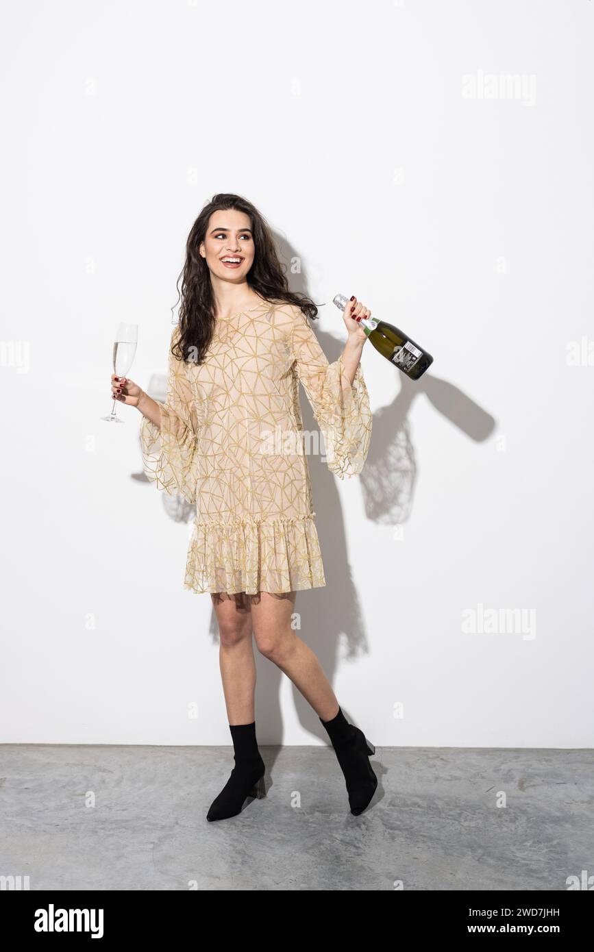Joyeuse femme élégante s'amusant à la fête, célébrant quelque chose, dansant avec une bouteille et un verre de champagne, debout sur fond blanc Banque D'Images