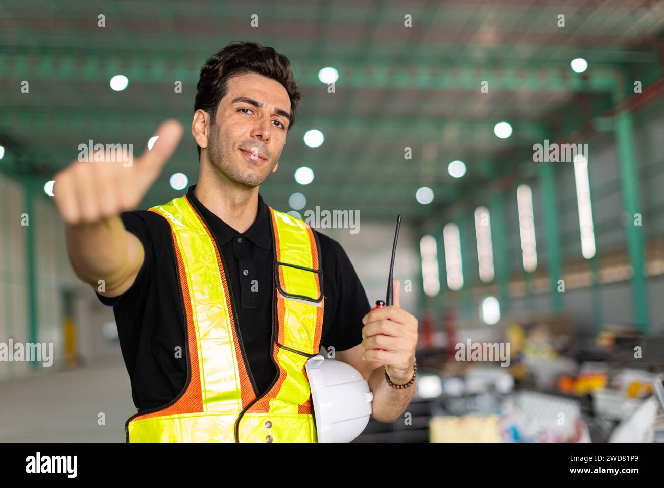 La main de l'homme d'ingénieur comme signe ou ouvrier d'usine portent le casque de sécurité dans le site d'entrepôt intérieur montrant les pouces vers le haut sourire heureux. Bon travail terminé fait Banque D'Images