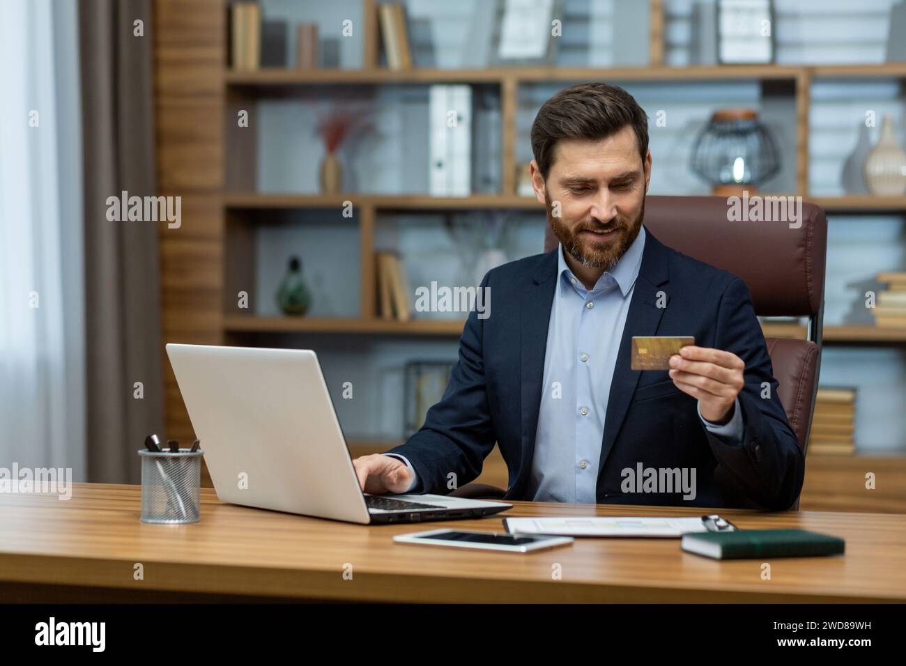 Homme d'affaires mature dans le bureau à domicile tenant la carte de crédit, envisageant le paiement en ligne ou l'investissement sur ordinateur portable. Banque D'Images