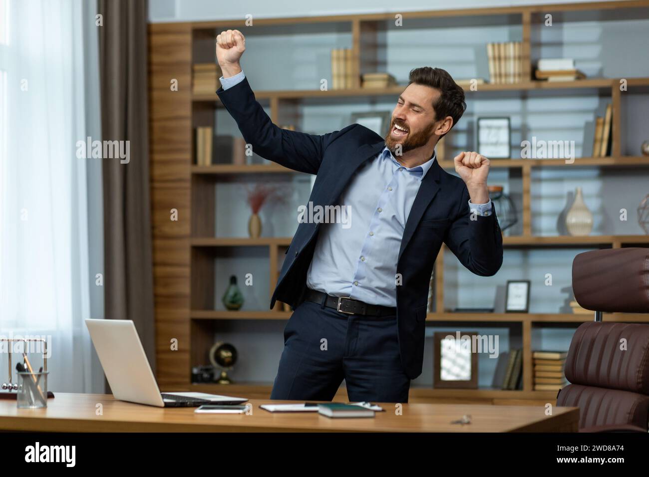 Homme d'affaires mature énergique dans un costume intelligent célèbre la victoire avec une pompe de poing dans un environnement de bureau à domicile bien aménagé. Banque D'Images