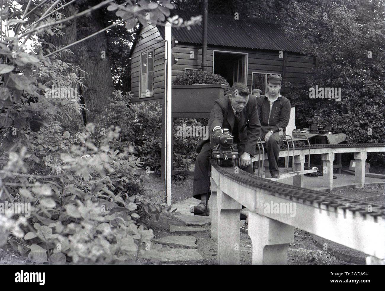 Années 1950, historique, à l'extérieur dans un jardin, amateurs de chemin de fer miniature dans un chemin de fer à vapeur de jardin miniature, à la Field End Railway Co, Angleterre, Royaume-Uni. Sur une voie surélevée assise sur des poteaux en béton, deux hommes montent sur de minuscules trains à vapeur, l'un étant un modèle réduit de la locomotive à vapeur GWR 'County of Oxford' (1023), dont l'original a commencé à fonctionner en 1947. Banque D'Images