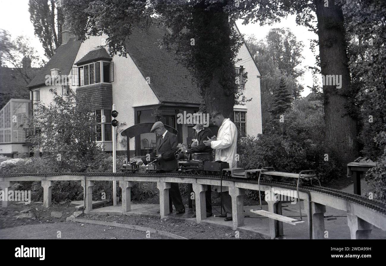 Années 1950, historique, à l'extérieur dans un jardin d'une grande maison de campagne, amateurs de chemin de fer modèle, un dans un manteau blanc, à côté d'une voie de chemin de fer à vapeur de jardin miniature, à la Field End Railway Co, Angleterre, Royaume-Uni. Sur une voie surélevée reposant sur des poteaux en béton, les trains à vapeur miniatures, l'un étant un modèle réduit de la locomotive à vapeur GWR 'County of Oxford' (1023), dont l'original a été mis en service en 1947. Banque D'Images