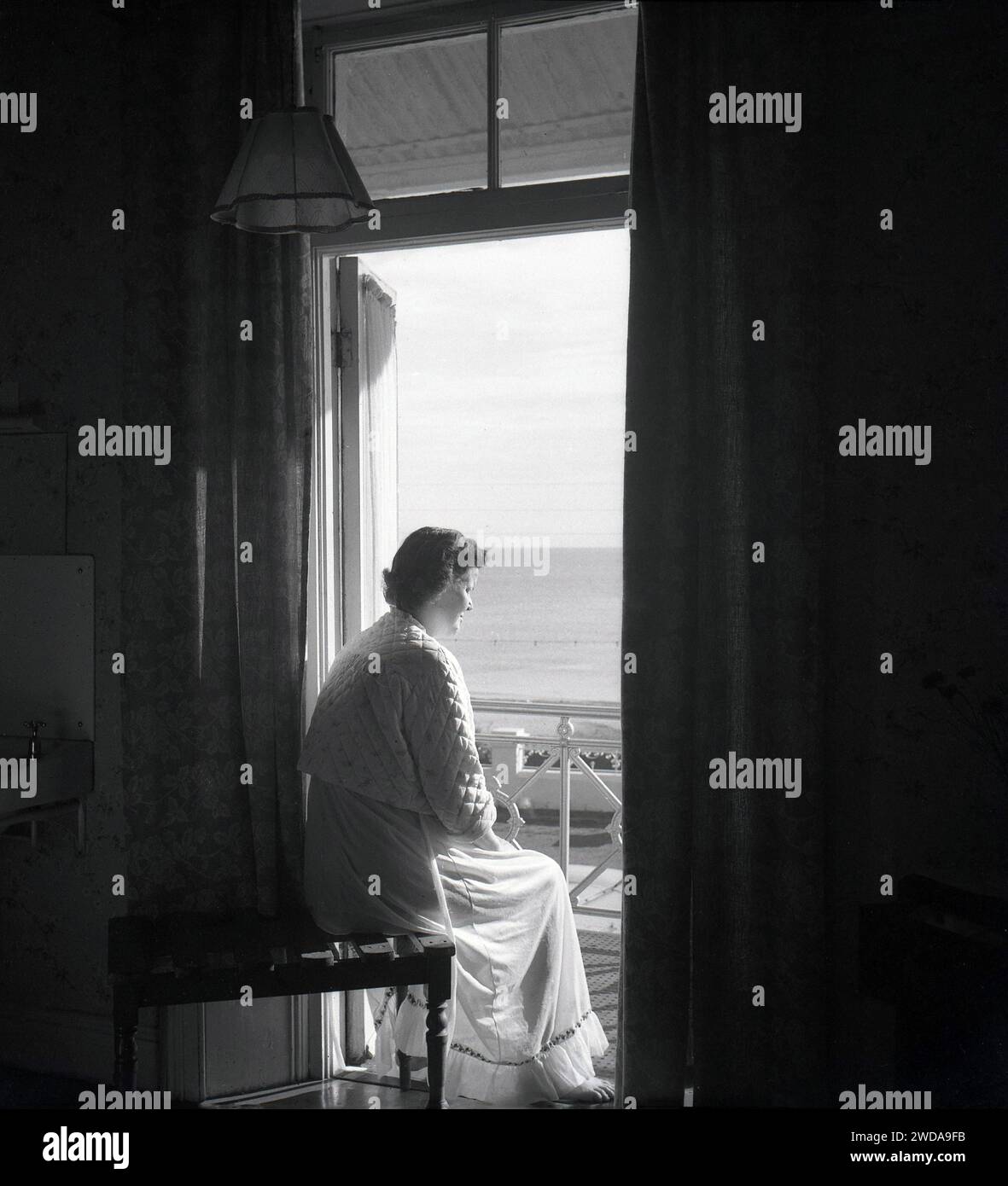Années 1950, historique, tôt le matin et une dame dans une chemise de nuit et un dessus de lit matelassé assis sur un banc à bagages en ardoise en bois à l'intérieur d'une chambre d'hôtel par son balcon donnant sur la mer, Angleterre, Royaume-Uni. Banque D'Images