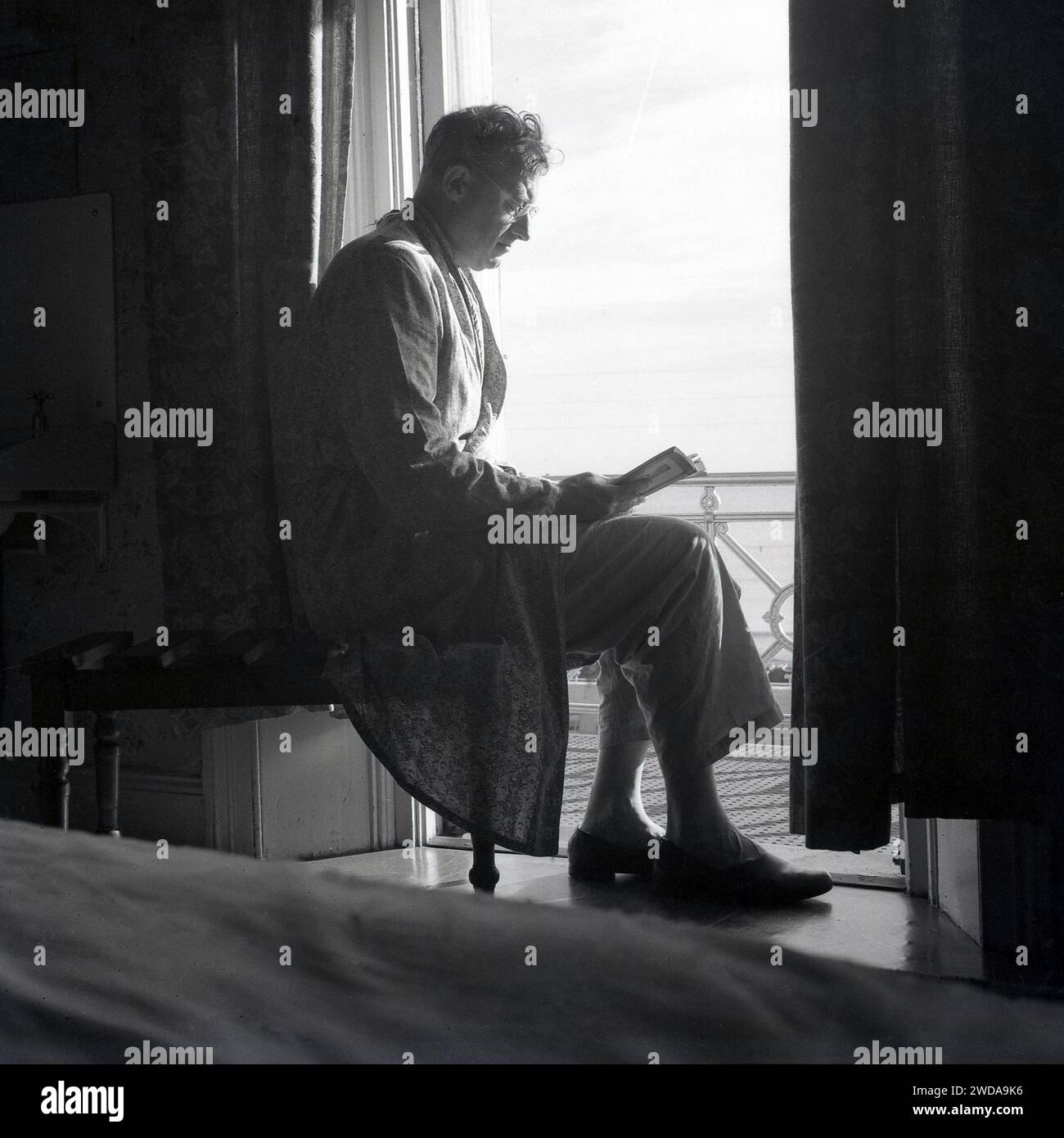 Années 1960, historique, à l'intérieur d'une chambre d'hôtel, tôt le matin et un gentleman en pyjama et une robe de chambre en coton léger assis lisant un livre par le balcon donnant sur la mer, Angleterre, Royaume-Uni. Banque D'Images