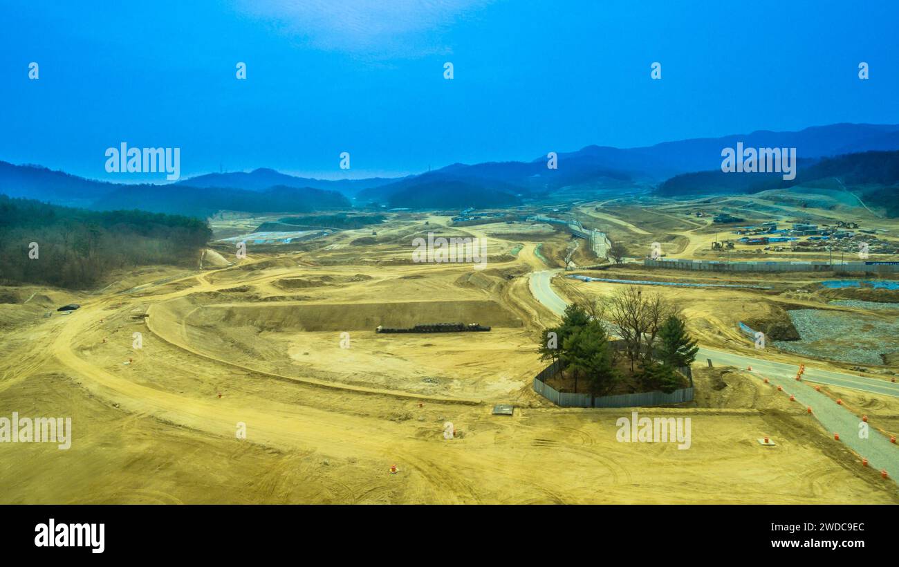 Vue aérienne du chantier de construction avec un petit groupe d'arbres clôturés pour la protection et des collines ondulantes sous le ciel bleu en arrière-plan, Corée du Sud Banque D'Images