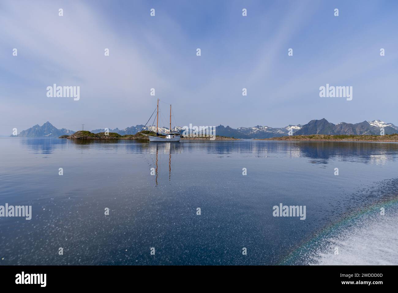 La mer calme reflète un voilier et des montagnes norvégiennes escarpées, avec un arc-en-ciel subtil des éclaboussures d'eau dans les paisibles Lofoten Banque D'Images
