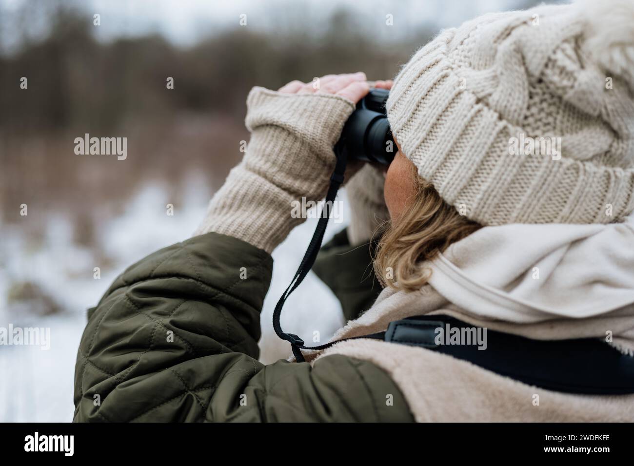 Femme vêtue de vêtements d'hiver avec des gants et un bonnet tricoté à la recherche d'oiseaux et d'animaux à travers les jumelles dans la nature Banque D'Images