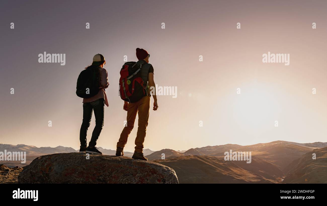 Deux jeunes silhouettes actives de randonneurs avec des sacs à dos sont debout sur un gros rocher et regardent le coucher du soleil dans les montagnes Banque D'Images