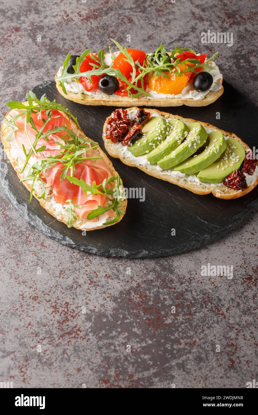Trois sandwichs ouverts différents avec roquette, jamon, tomates séchées au soleil, avocat, fromage à la crème, olives et poivrons rôtis gros plan sur une planche d'ardoise o Banque D'Images