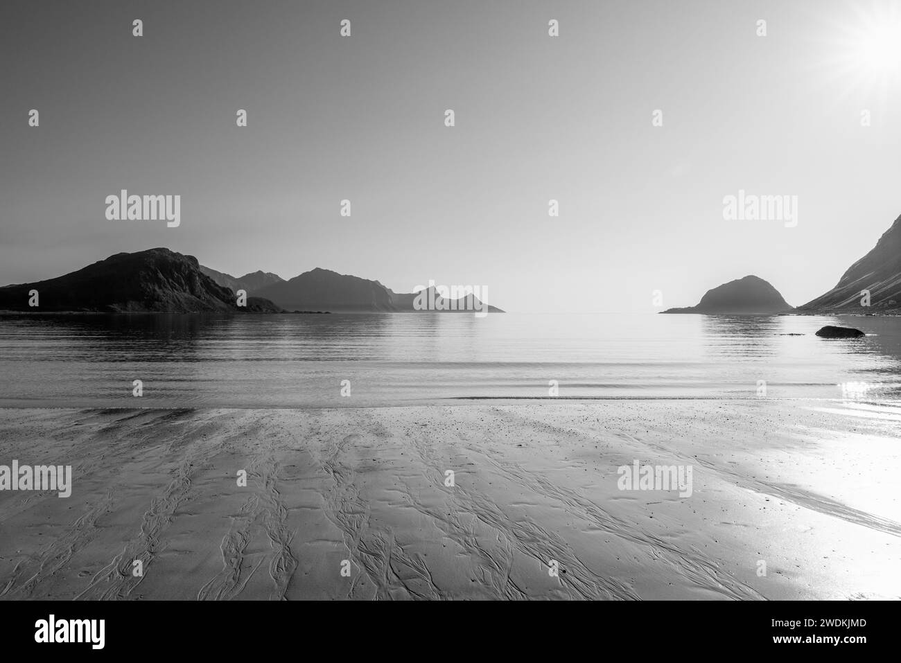 Image monochrome capturant les contrastes saisissants des textures de marée sur Haukland Beach, Lofoten avec le soleil suspendu au-dessus de la mer tranquille Banque D'Images