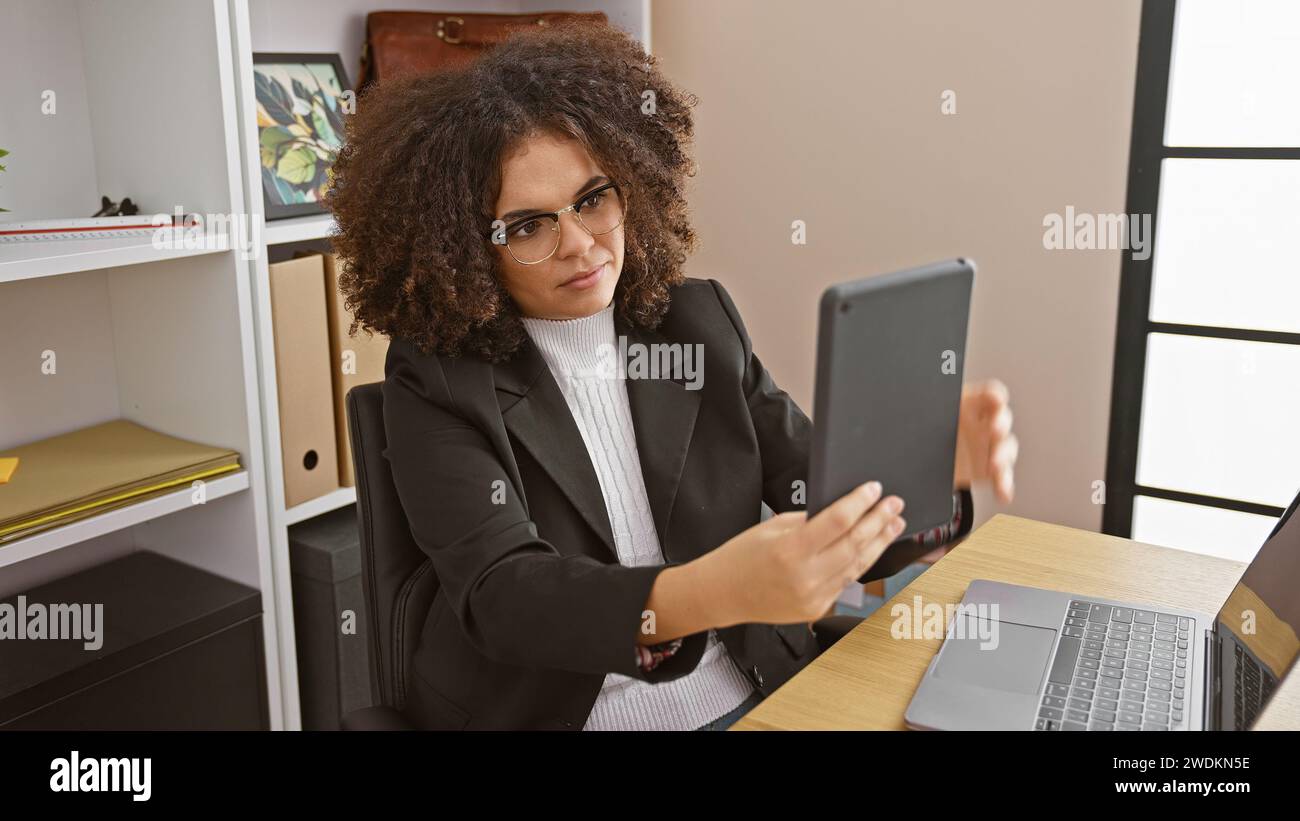 Une jeune femme hispanique aux cheveux bouclés portant des lunettes et un blazer utilise une tablette dans un bureau moderne. Banque D'Images